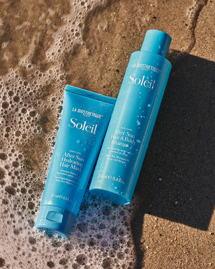 After Sun Hydrating Hairmask und After Sun Hair & Body Shampoo in hellblauer Verpackung mit weißer Schrift, vor Sand- und Meerwasser-Hintergrund – intensive Sommerpflege für Haut und Haar